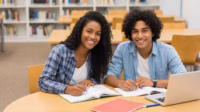 Two students sitting at a table in a library, smiling at the camera while writing in open textbooks, with a laptop and notebooks in front of them.